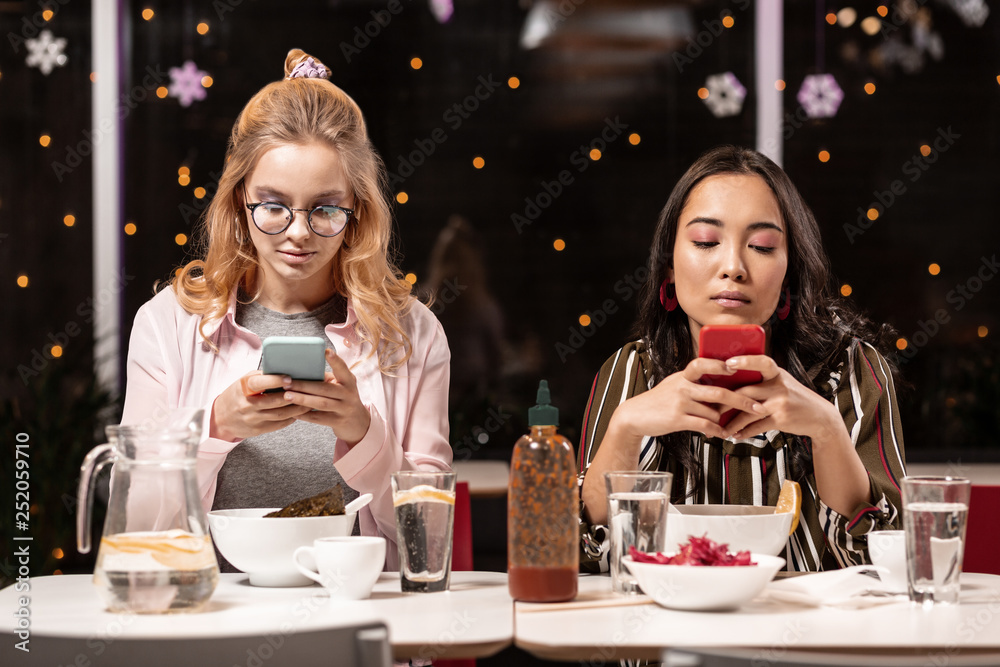 Two girls spending most of the time with their smartphones while sitting in restaurant.