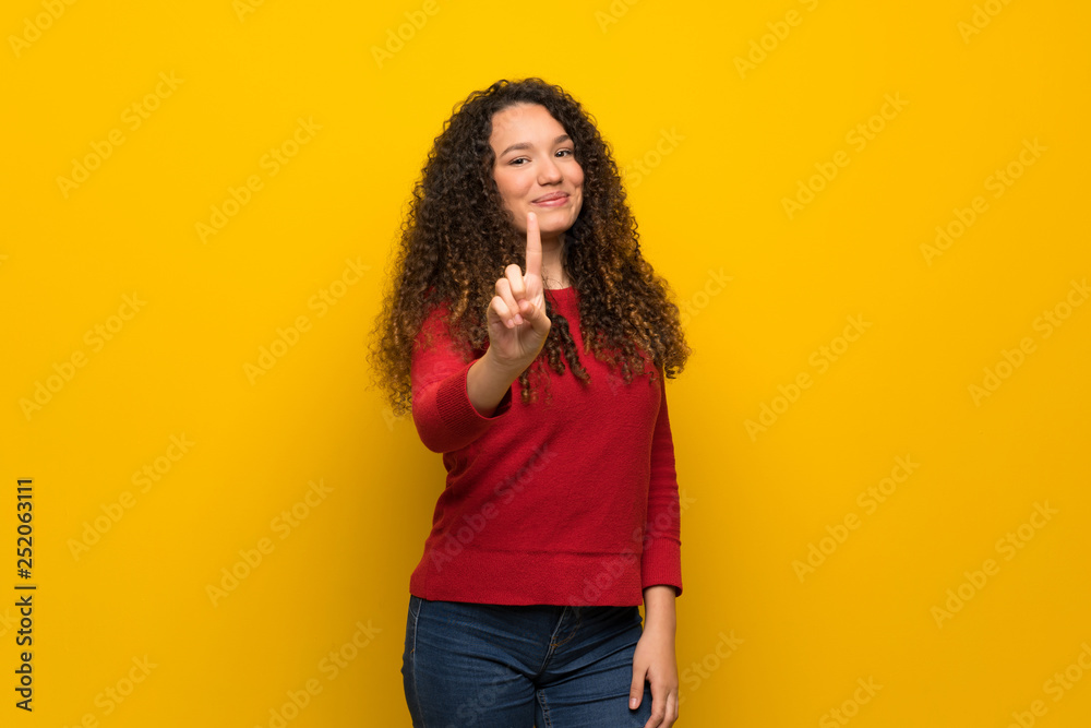 Teenager girl with red sweater over yellow wall showing and lifting a finger Teenager girl with red sweater over yellow wall showing and lifting a finger