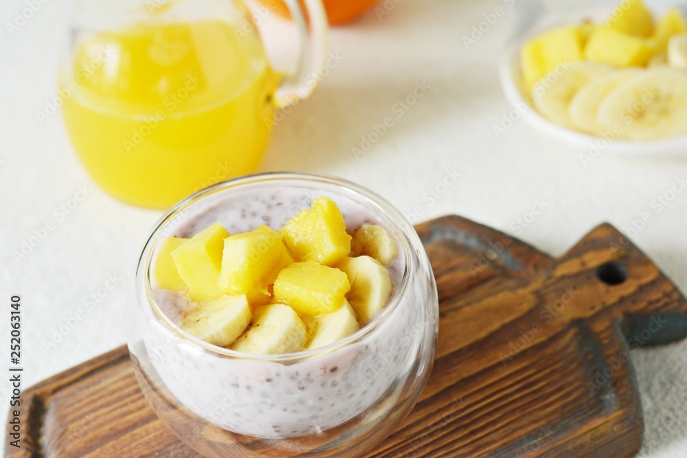 Chia pudding with mango pulp  in beautiful glasses with green mint leaves and cut fresh ripe tropical fruit, orange juice for breakfast on a light background - raw vegetarian sweet organic dessert. 