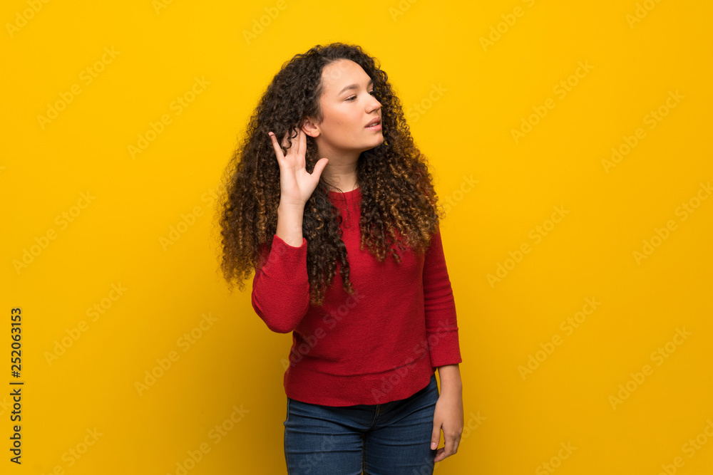 Teenager girl with red sweater over yellow wall listening to something by putting hand on the ear Teenager girl with red sweater over yellow wall listening to something by putting hand on the ear