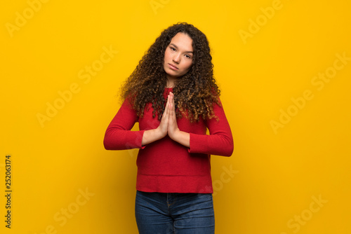 Teenager girl with red sweater over yellow wall keeps palm together. Person asks for something © luismolinero