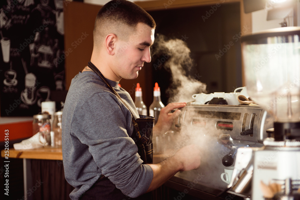 Professional barista holding metal jug warming milk using the coffee machine. Happy young man preparing coffee at counter Professional barista holding metal jug warming milk using the coffee machine. Happy young man preparing coffee at counter