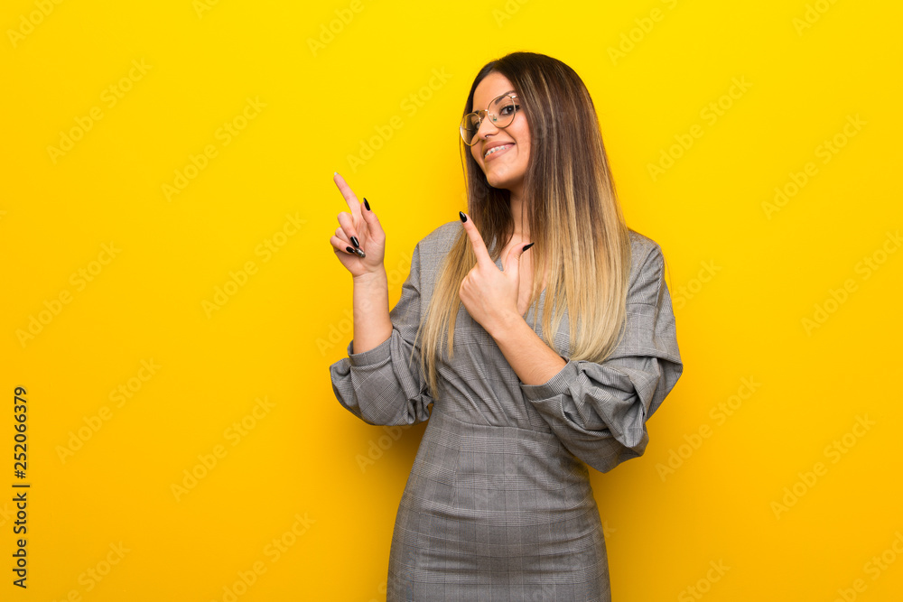 Young woman with glasses over yellow wall pointing with the index finger and looking up Young woman with glasses over yellow wall pointing with the index finger and looking up