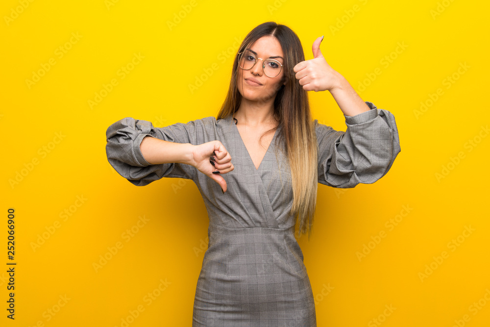Young woman with glasses over yellow wall making good-bad sign. Undecided between yes or not Young woman with glasses over yellow wall making good-bad sign. Undecided between yes or not