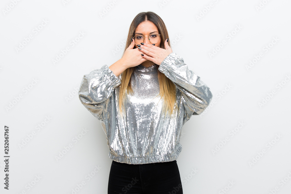 Young woman with glasses over white wall covering mouth with hands for saying something inappropriate