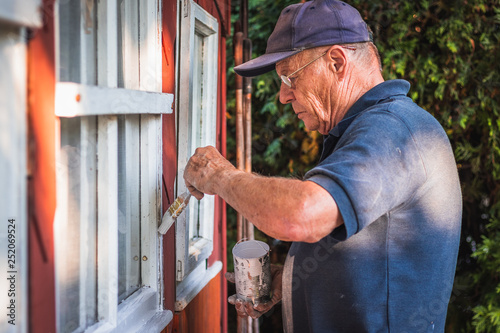 Fotografi Active senior man painting window frame of wooden cabin
