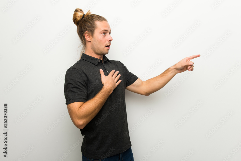 Blond man with long hair over white wall pointing finger to the side with a surprised face Blond man with long hair over white wall pointing finger to the side with a surprised face