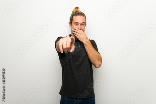 Blond man with long hair over white wall pointing with finger at someone and laughing