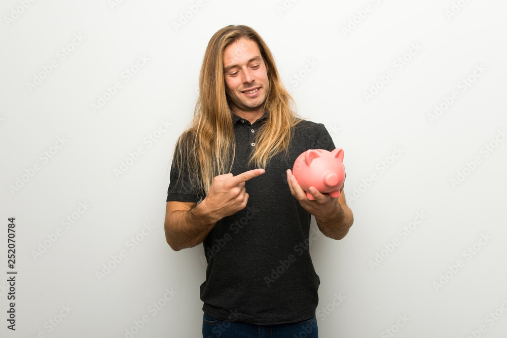 Blond man with long hair over white wall holding a piggybank