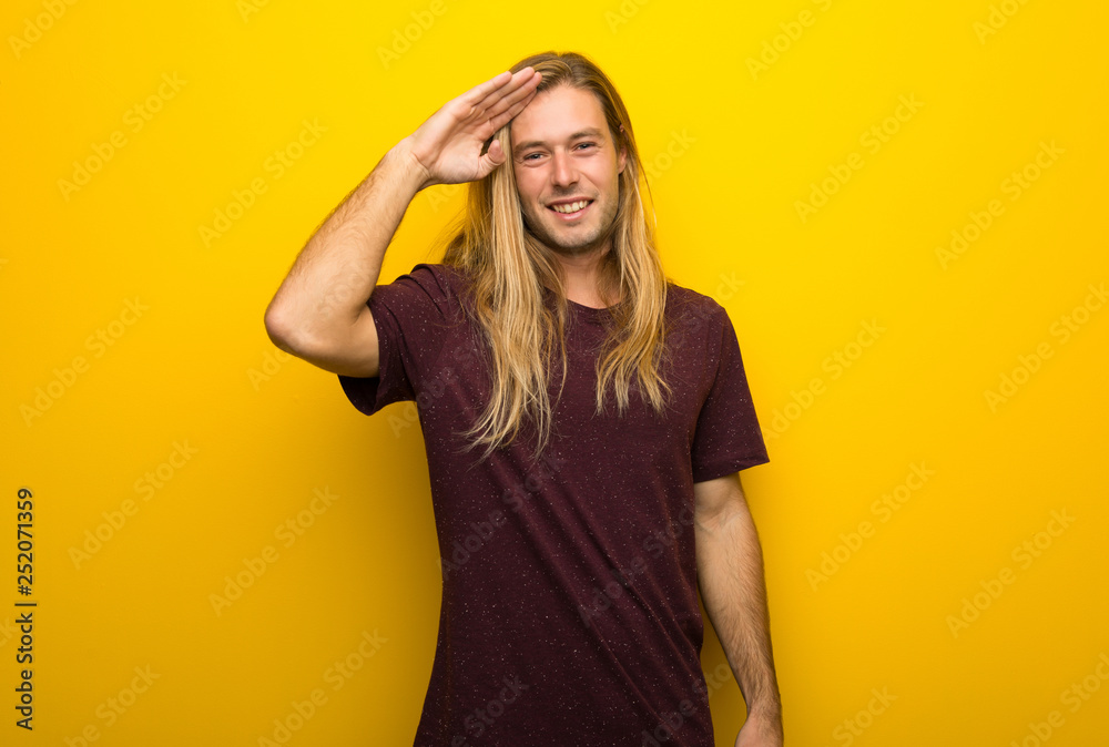 Blond man with long hair over yellow wall saluting with hand Blond man with long hair over yellow wall saluting with hand
