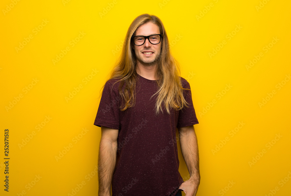 Blond man with long hair over yellow wall with glasses and happy