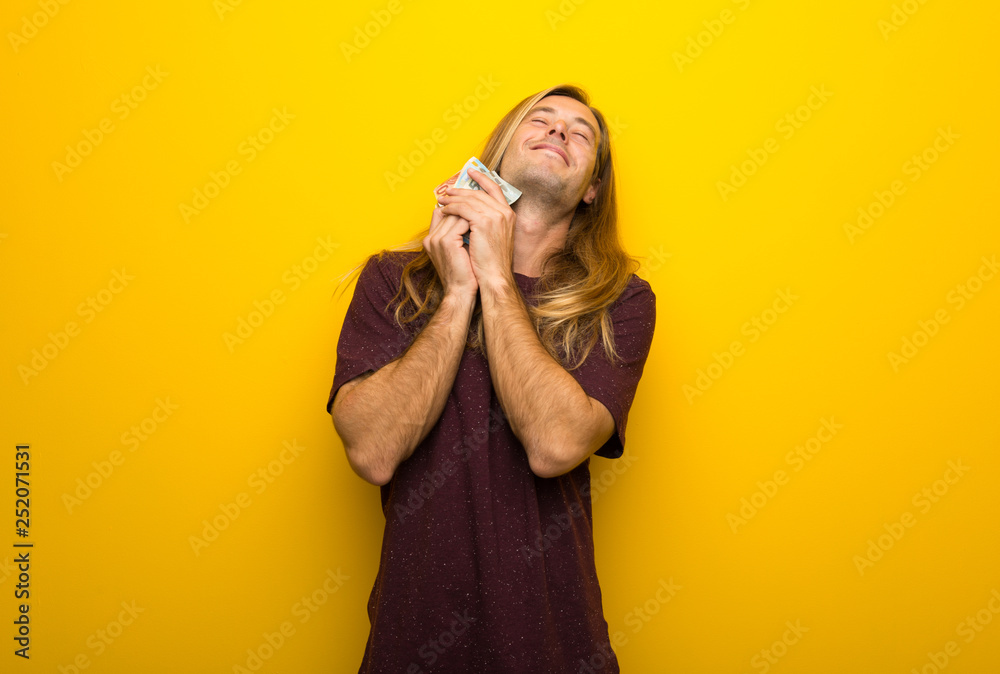 Blond man with long hair over yellow wall taking a lot of money Blond man with long hair over yellow wall taking a lot of money