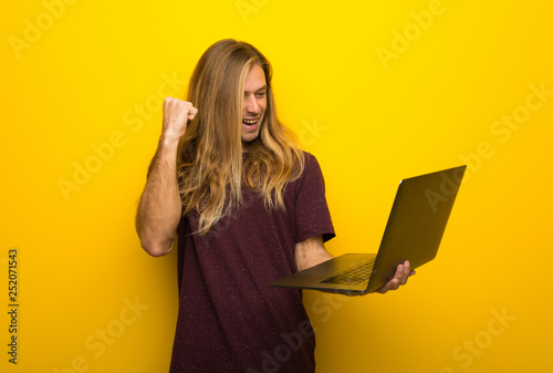 Blond man with long hair over yellow wall with laptop and celebrating a victory