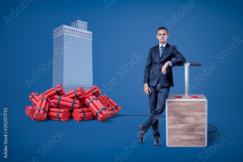 Fototapeta Businessman standing next to detonator box connected to heap of dynamite beside high-rise building in the background