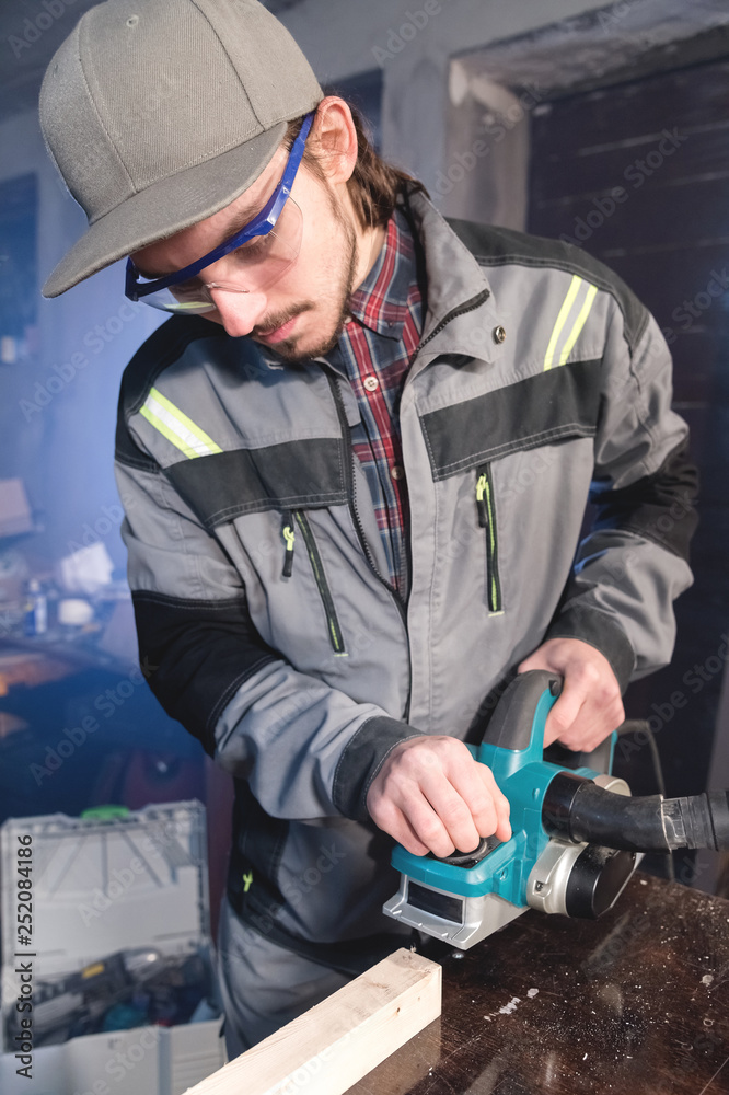 Young carpenter with a beard working with an electric plane with suction of sawdust. Leveling and sanding wooden bars Young carpenter with a beard working with an electric plane with suction of sawdust. Leveling and sanding wooden bars