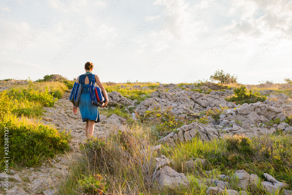 Young woman walking on a rocky path carrying beach blankets and beach head rest wearing a blue dress. Summer holidays on island Krk in Croatia. Travel and summer concept.