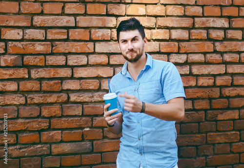 Portrait of a dissatisfied or surprised guy, holding a coffee in his hand against a brick wall, wearing a blue shirt, urban style
