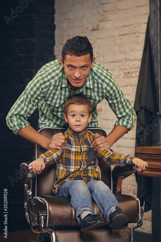 stylish little kid sitting on chair at barbershop with his young father on background