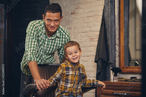 stylish little kid sitting on chair at barbershop with his young father on background
