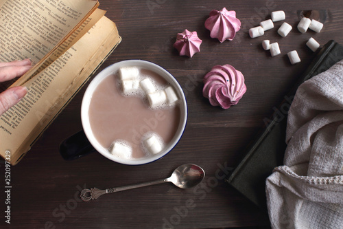 Сocoa coffe with marshmallows in a dark blue cup on a wooden table next to a book, a spoon and meringue top view atmospheric cozy flat lay