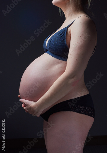 A beautiful pregnant woman body, with the woman gently holding and supporting the pregnant stomach. Gentle feminine motherhood. Photographed on a black studio background.