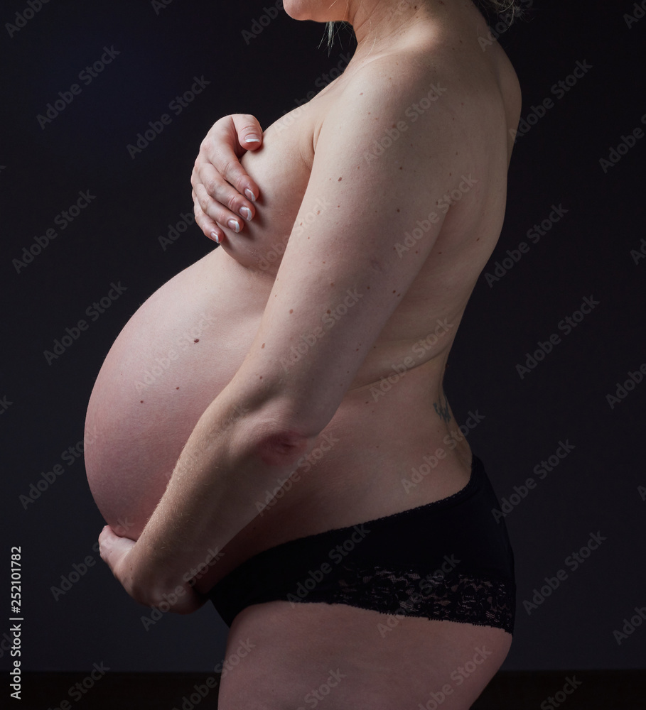 Naklejka premium A beautiful pregnant woman body, with the woman gently holding and supporting the pregnant stomach. Gentle feminine motherhood. Photographed on a black studio background.