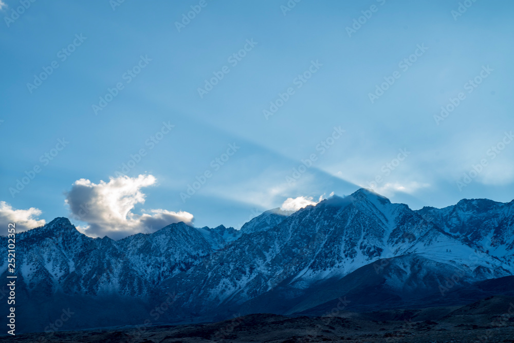 blue sky, clouds, slanted beams of sunlight over peaks of Eastern Sierra Nevada, California, USA