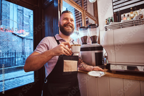 Cheerful barista in apron holding a cup and saucer, drinking coffee during lunch break leaning on a counter in the coffee shop.