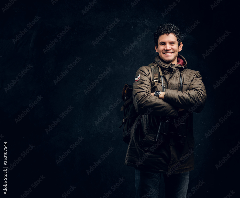 Confident young traveller in hiking gear with binoculars stands with crossed hands, smiling and looking at a camera in the studio against a dark wall Confident young traveller in hiking gear with binoculars stands with crossed hands, smiling and looking at a camera in the studio against a dark wall