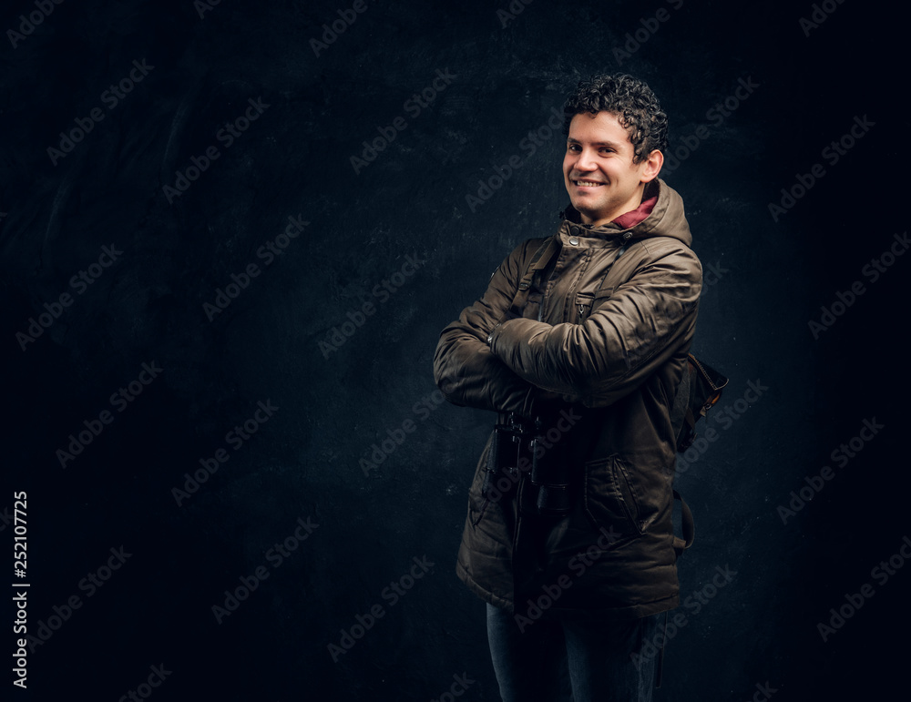 Confident young traveller in hiking gear with binoculars stands with crossed hands, smiling and looking at a camera in the studio against a dark wall Confident young traveller in hiking gear with binoculars stands with crossed hands, smiling and looking at a camera in the studio against a dark wall