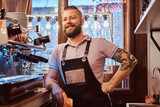Confident barista with stylish beard and hairstyle wearing apron smiling and looking sideways while leaning on a counter in the cafe or restaurant