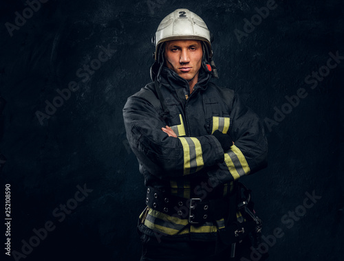 Brutal fireman in uniform posing for the camera standing with crossed arms and confident look. Studio photo against a dark textured wall