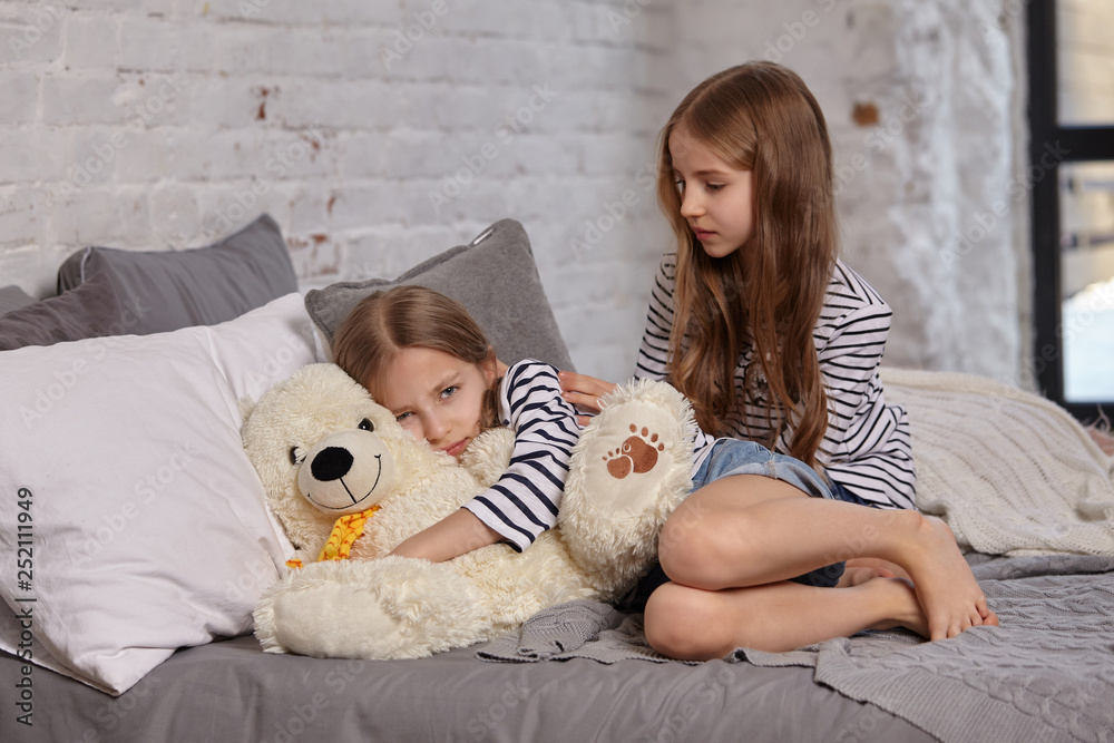 The image of two little sisters sitting on the bed in the room. The image of two little sisters sitting on the bed in the room.