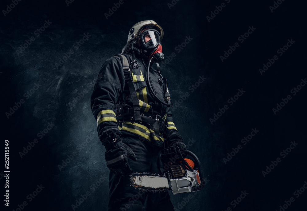 Firefighter in safety helmet and oxygen mask holding a chainsaw. Studio photo against a dark textured wall