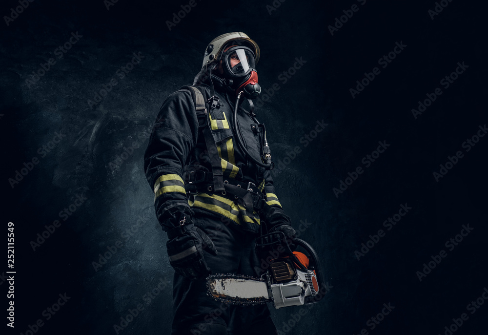 Firefighter in safety helmet and oxygen mask holding a chainsaw. Studio photo against a dark textured wall Firefighter in safety helmet and oxygen mask holding a chainsaw. Studio photo against a dark textured wall