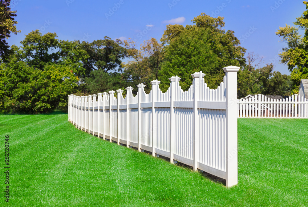 White vinyl fencing spanning across a back yard with green grrass, trees and blue sky in the background White vinyl fencing spanning across a back yard with green grrass, trees and blue sky in the background