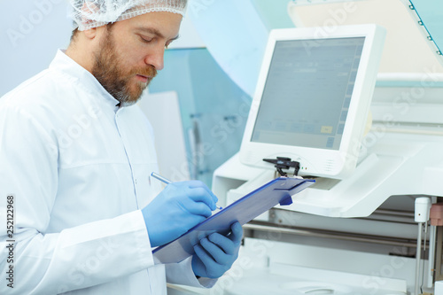 Side view of male laboratory assistant in white cap and blue gloves doing experiments. Attractive doctor with red beard in lab coat checking new modern equipment. Medical worker writing results.