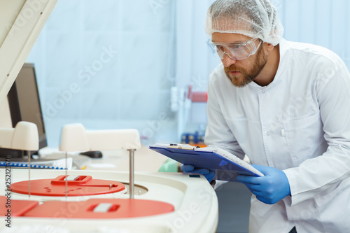 Young man in glasses and cap doing experiment in laboratory. Handsome doctor with red beard writing results of research. Medical worker in lab coat and blue gloves checking new modern equipment.