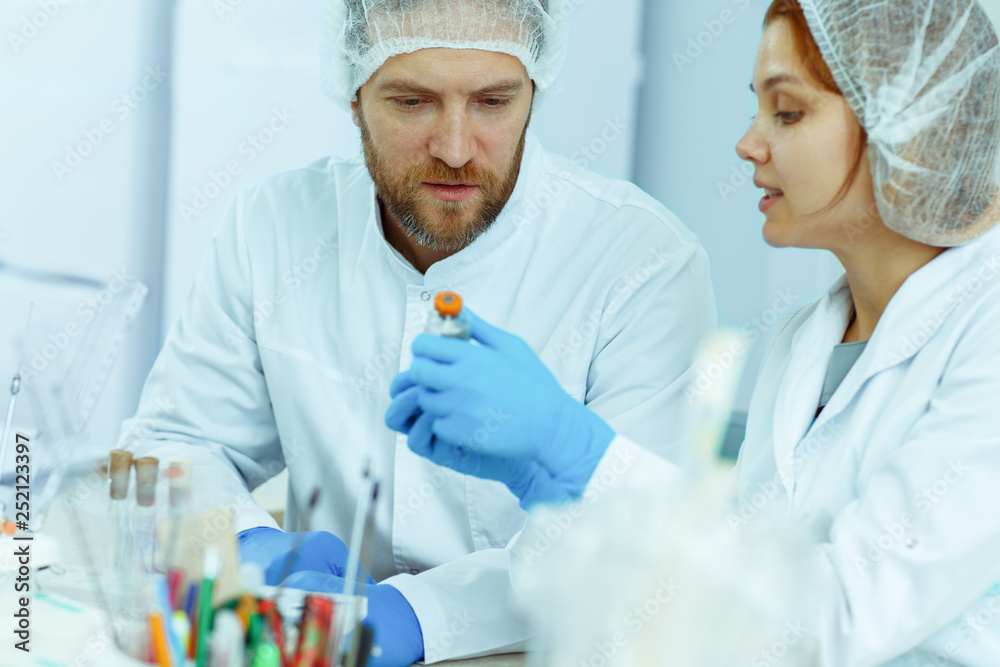 Beautiful red haired woman in lab coat holding test tube in her hand. Medical workers sitting in modern laboratory during work. Young scientist doing research and talking with her colleague. Beautiful red haired woman in lab coat holding test tube in her hand. Medical workers sitting in modern laboratory during work. Young scientist doing research and talking with her colleague.