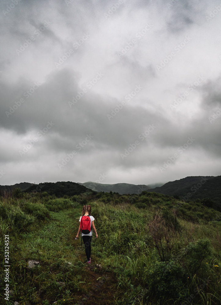 A little girl putnix red backpack goes along the path among the green hills. Cloudy weather.