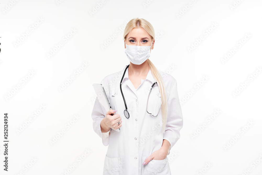 Portrait of young female blond doctor in medical mask and white working uniform looking at camera isolated on white. A stethoscope is hanging on the neck Portrait of young female blond doctor in medical mask and white working uniform looking at camera isolated on white. A stethoscope is hanging on the neck