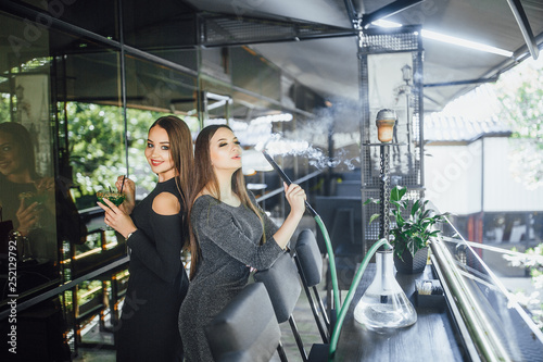 Two beautiful girls are standing on the summer terrace of a modern cafe and smoking hookah