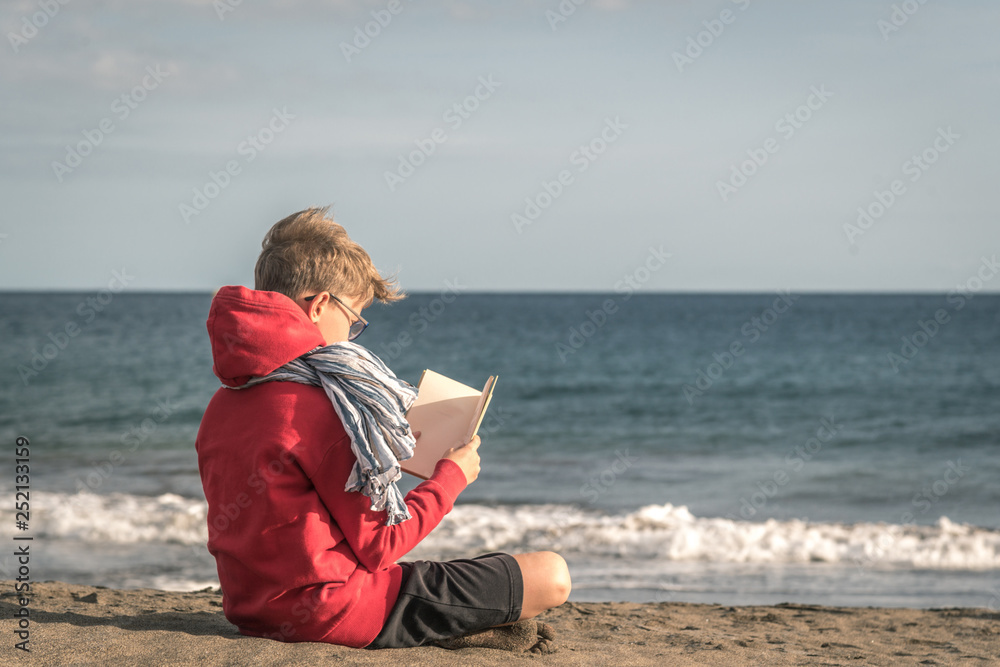 Fototapeta premium Cute boy in a red hoodie reading a book at the beach in a cloudy day, enjoying holiday. Young male sitting on the sand and relaxing with a blast. Kid enjoying a trip in a new exotic place near the sea