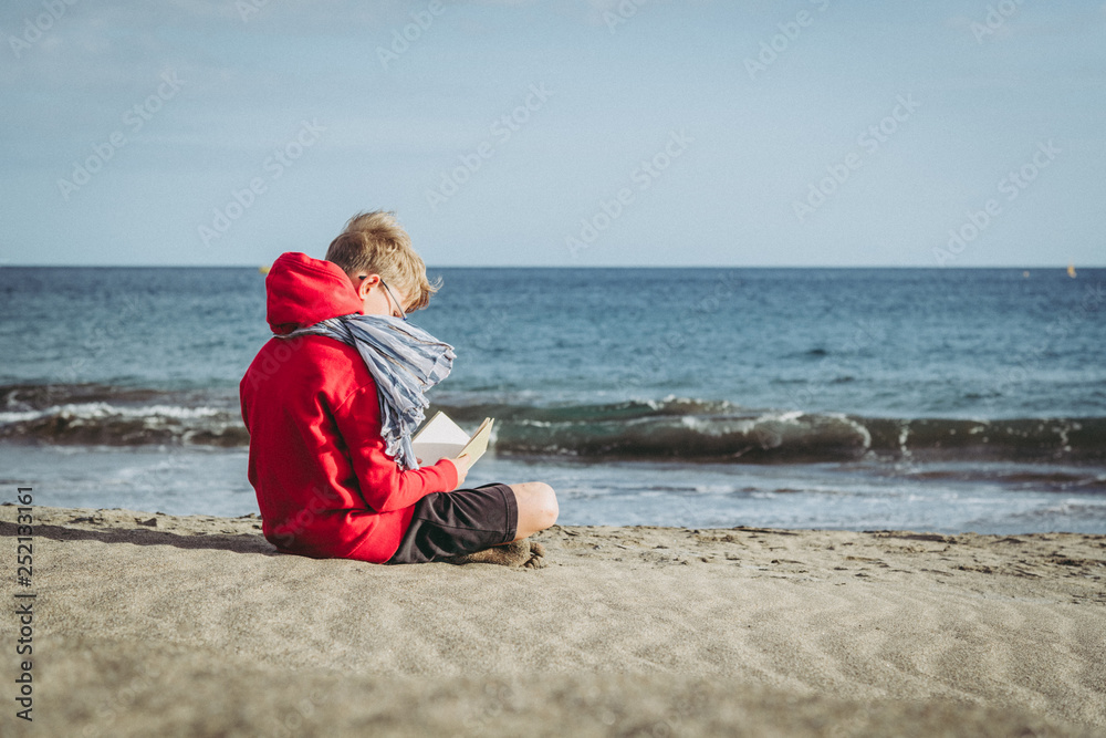 Cute boy in a red hoodie reading a book at the beach in a cloudy day, enjoying holiday. Young male sitting on the sand and relaxing with a blast. Kid enjoying a trip in a new exotic place near the sea