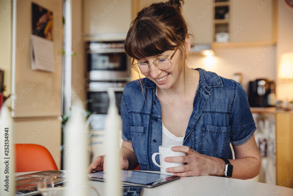Smiling woman sitting with cup of coffee at table in the kitchen using tablet Smiling woman sitting with cup of coffee at table in the kitchen using tablet