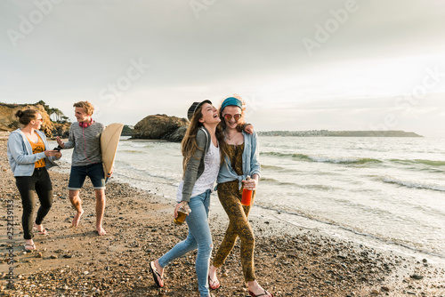 Happy friends with surfboard and drinks walking on stony beach photo