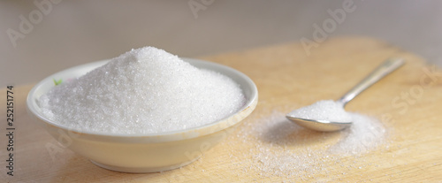 Sugar in a white cup with a spoon with white sugar placed on the side of a cup on a wooden floor.
