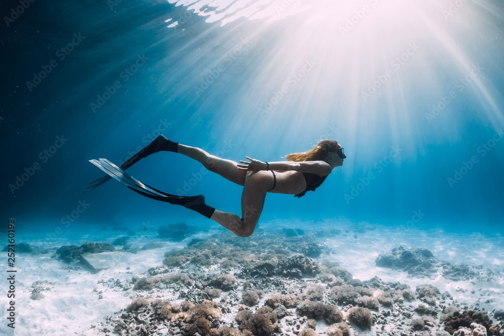 Pretty woman freediver with fins swim over sandy sea and sun rays underwater