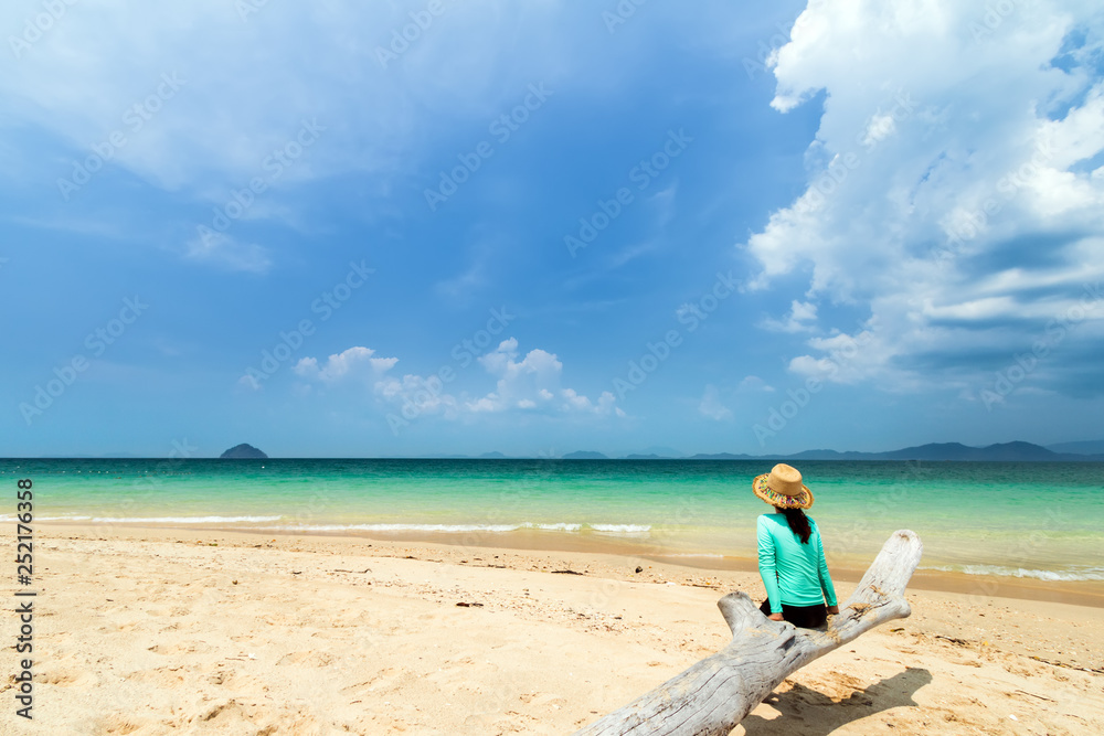 Young beautiful woman wear hat weave in swimsuit sitting on tree timber over white sand beach of andaman sea at summer sunny day