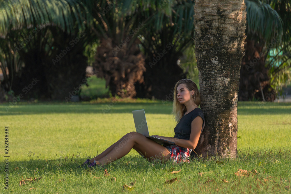 young beautiful woman in black top with laptop on her knees sits under palm